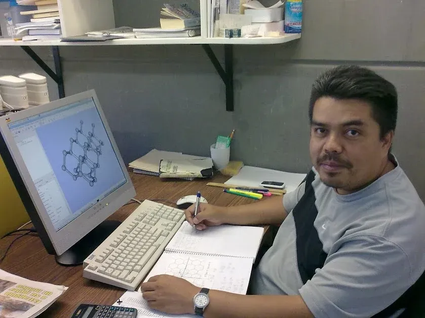 El Dr. Cabellos, durante su doctorado, escribe un análisis estructural a mano junto a un monitor que muestra un modelo molecular.