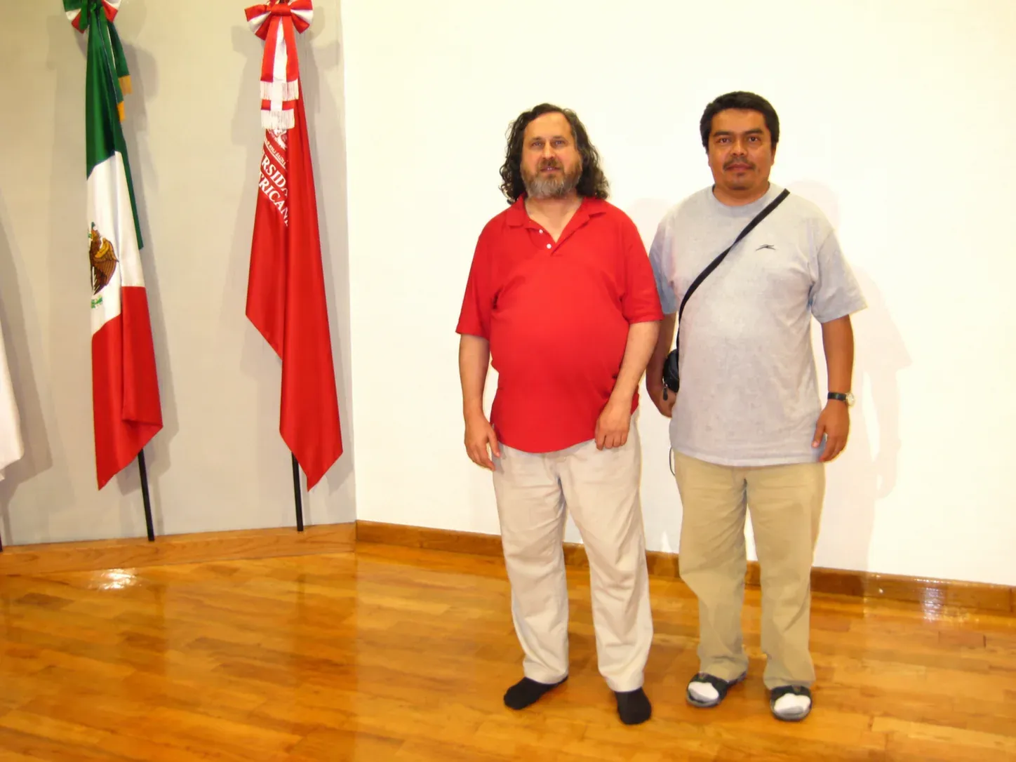 Richard Stallman, con camisa roja, de pie junto al Dr. José Luis Cabellos en un salón de la Universidad Iberoamericana León, con banderas al fondo.